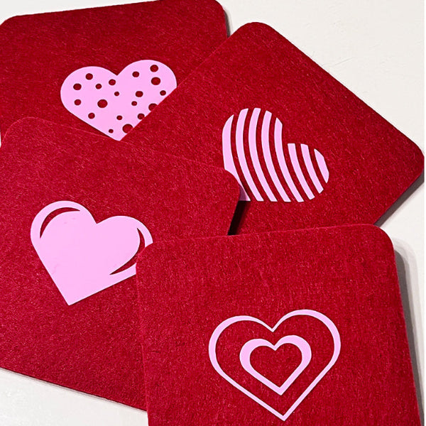 Closeup product image of a set of four square shaped wool red drink coasters with pink heart heat transfer designs on a white table