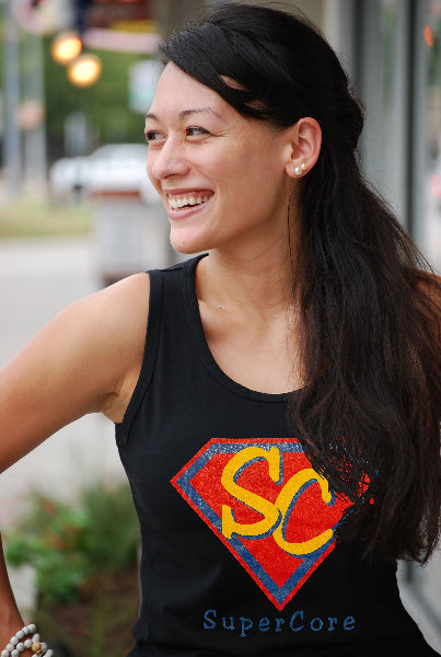 Image of a woman outside of a building, wearing a black tank top with a red graphic and the word super core printed in blue