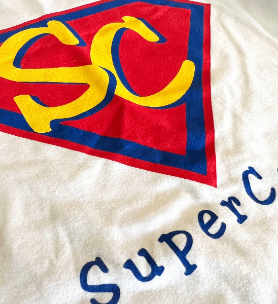 Closeup detail product image of a printed graphic on a white knit fabric t-shirt. Graphic is a red and blue diamond with the letter s and c printed inside the diamond 