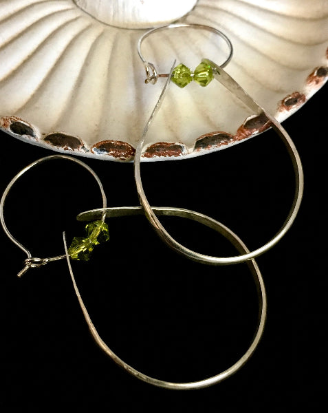 Closeup image of a pair of hammered sterling silver double hoop earrings with green swarovski crystals laying partially in a shell shaped dish