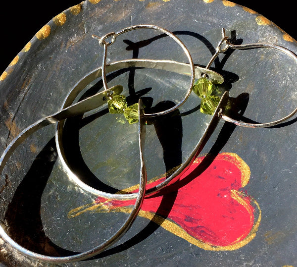 Closeup image of a pair of hammered sterling silver double hoop earrings with green swarovski crystals laying inside of a painted dish with a painted heart