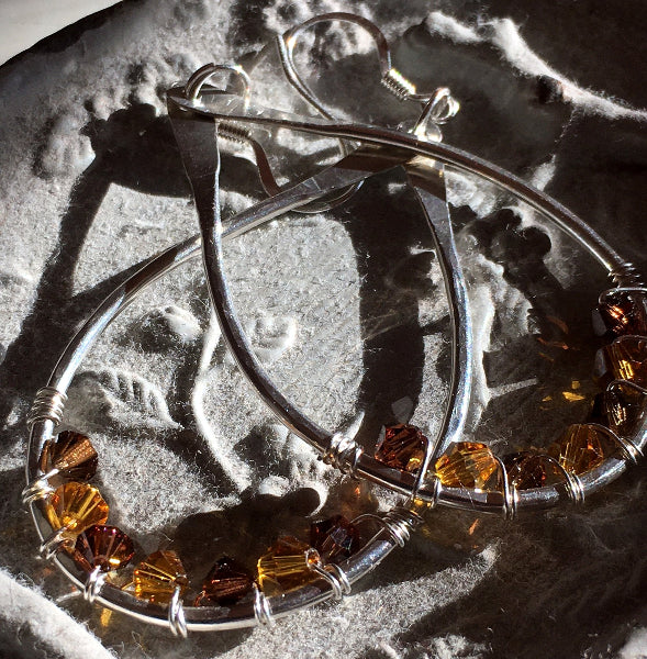 Closeup image of a pair of hammered sterling silver teardrop hoop earrings with silver wire wrap and swarovski crystals