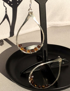 Closeup image of a pair of hammered sterling silver teardrop hoop earrings with silver wire wrap and swarovski crystals hanging from and laying in a black earring tree stand on a table