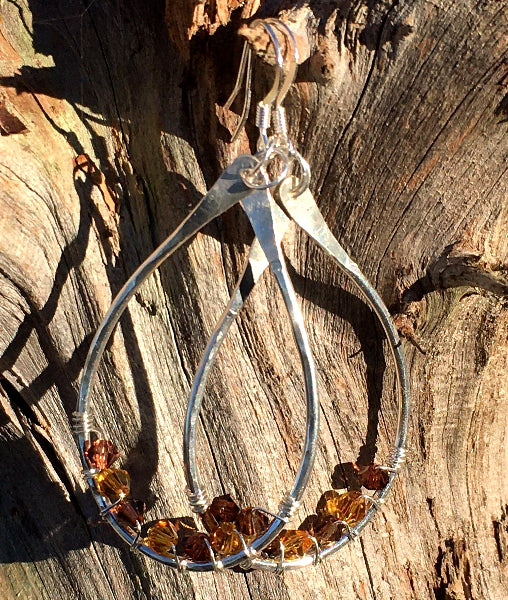 Closeup image of a pair of hammered sterling silver teardrop hoop earrings with silver wire wrap and swarovski crystals hanging from a tree stump in the sun