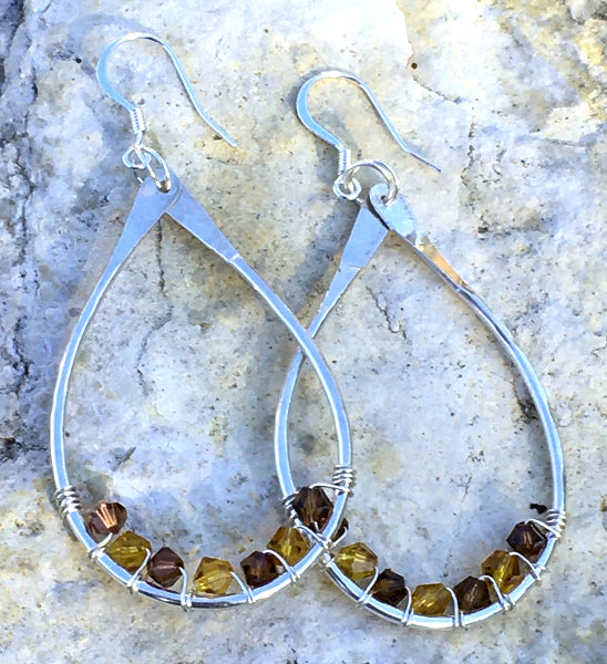 Closeup image of a pair of hammered sterling silver teardrop hoop earrings with silver wire wrap and swarovski crystals laying on a stone 