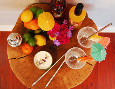 Top view image of a wood table with a bowl of citrus fruit, bottles of alcohol beverages, small olive bowl and flowers and two clear glasses with paper drink umbrellas and stainless-steel metal straws