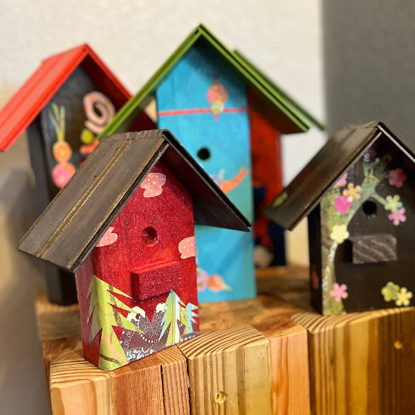 Product image of decorative birdhouses with decoupaged whimsical designs and wooden roof son top of a wooden stand