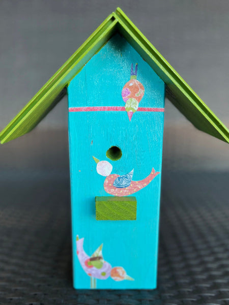 Closeup front view product image of aqua turquoise color decorative birdhouse with decoupaged whimsical bird design and green wooden roof on top of a wicker chair