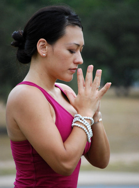 Image of a woman practicing yoga in a park setting, wearing a wine color yoga tank top, pearl earrings and a pearl bracelet