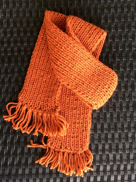 Image of a hand knit burnt orange child's scarf laying on black wicker 