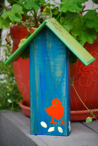 Back view of a hand painted blue decorative birdhouse with an orange chick painted at the bottom and a green roof, in front of a potted plant