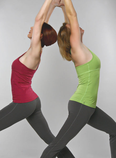 Image of two women practicing yoga poses standing back to back, wearing wine and green colored tank tops and grey boot cut yoga pants