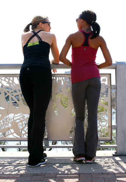 Back view of two women standing in front of an ornate fence. One woman is wearing black boot cut yoga pants, a black cross back tank op and black sneakers. The other woman is wearing grey boto cut yoga pants, a sangria pink color tank top and black sneakers