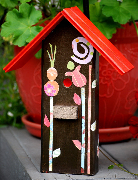 Closeup front view of a decorative wooden birdhouse with a red roof and decoupage floral design in front if a potted plant