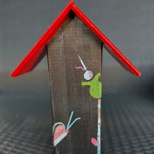 Back view of a decorative wooden birdhouse with a red roof and floral decoupaged design on a black wicker chair