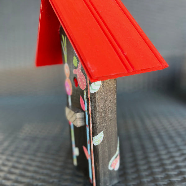 Side view of a brown metallic decorative wooden birdhouse with a red roof and decoupaged floral design on a black wicker chair