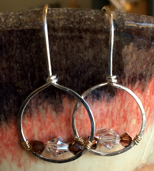 Closeup image of a pair of forward-facing hammered sterling silver hoop earrings with gold wire wrap and swarovski crystals hanging from the side of a ceramic bowl