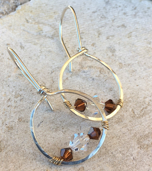 Closeup image of a pair of hammered sterling silver gold wire wrapped forward-facing hoop earrings with swarovski crystals displayed on cement background