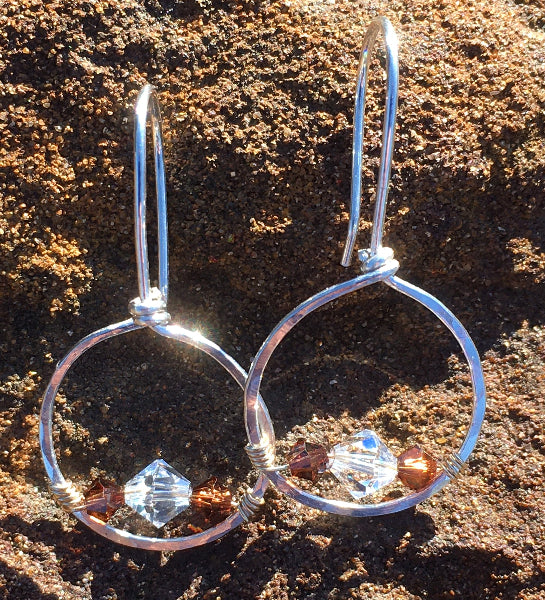 Closeup image of a pair of hammered sterling silver forward-facing hoop earrings with gold wire wrap and swarovski crystals on a rustic brown background 