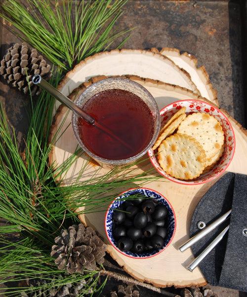 Top view of wood serving boards with pine needles and pinecones and a drink with a stainless-steel straw, bowl with black olives, a bowl with crackers and a charcoal wool straw travel case with stainless-steel straw