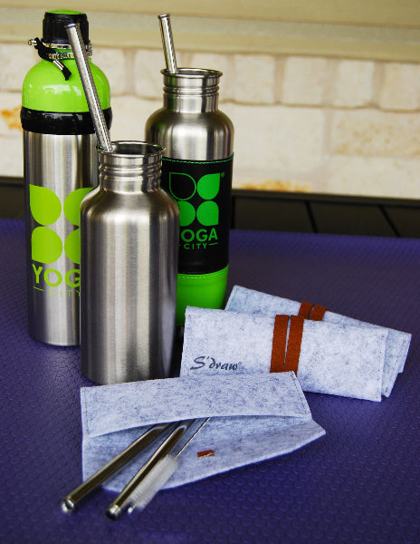 Image of metal water bottles with stainless-steel straws inside of them, standing on top of a purple yoga mat and light heather wool straw travel cases with faux leather straps