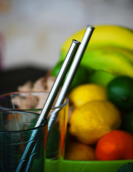 Closeup image of two stainless-steel straws inside of clear glasses with a bowl of fruit in the background