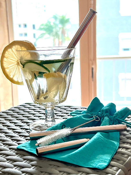 Image of a clear glass of cucumber lemon water with a stainless-steel drinking straw standing on top of a black wicker table in front of a window. The table has a stainless-steel metal extendable straw on top of an aqua colored drawstring travel bag with a wire handled straw cleaning brush