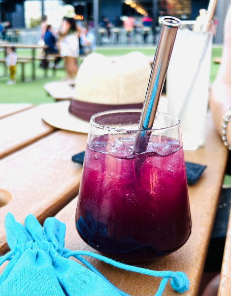 Closeup image of a clear glass of sangria on a wooden table with a stainless-steel straw and a turquoise aqua color drawstring travel bag. The  
 table is at a brewery in Austin, TX