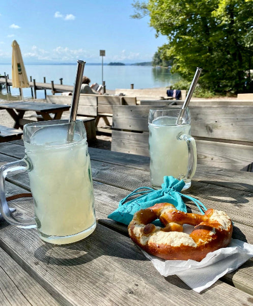 Image of a wooden picnic table and bench with a Bavarian pretzel and two glass steins with a citrus drink in them and stainless-steel metal extendable travel straws with a lakeview background