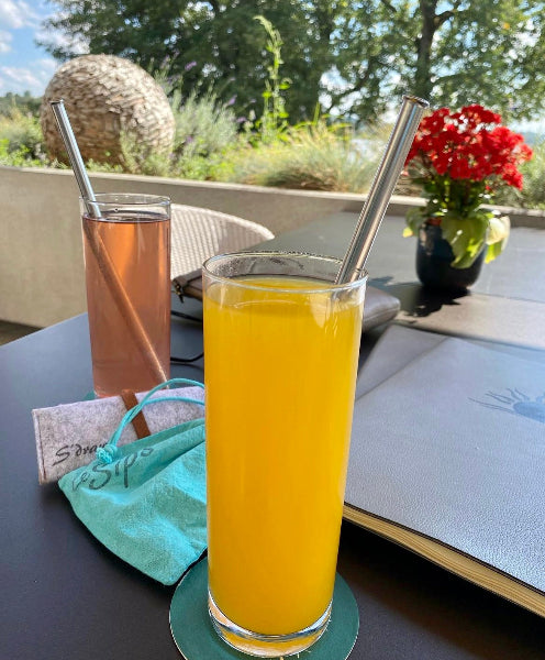 Image of two clear glasses with fruit drinks and long stainless-steel metal straws on top of coasters on a table in a garden setting 