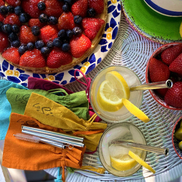 Top view image of a white wicker table with a berry tort on a plate, bowl with strawberries, two glasses of lemon ice water with sliced lemons and stainless-steel travel straws. Laying on the table is a stainless-steel extendable straw with a wire handle straw cleaning brush and colorful hand dyed drawstring bags