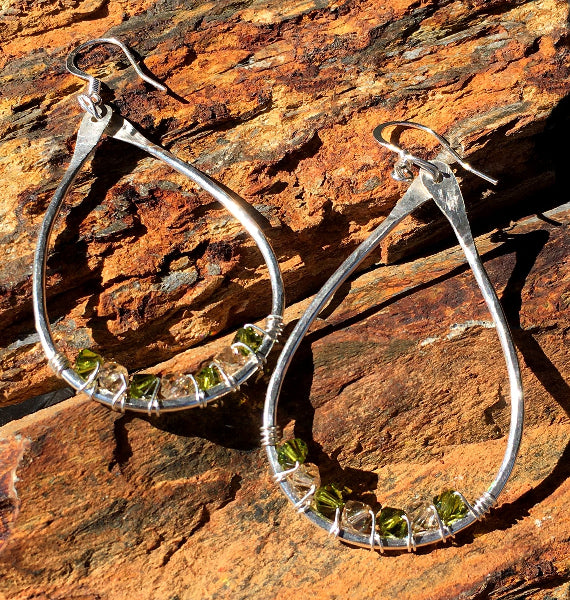 Closeup image of a pair of hammered sterling silver teardrop hoop earrings with silver wire wrap and swarovski crystals on petrified wood on the sun