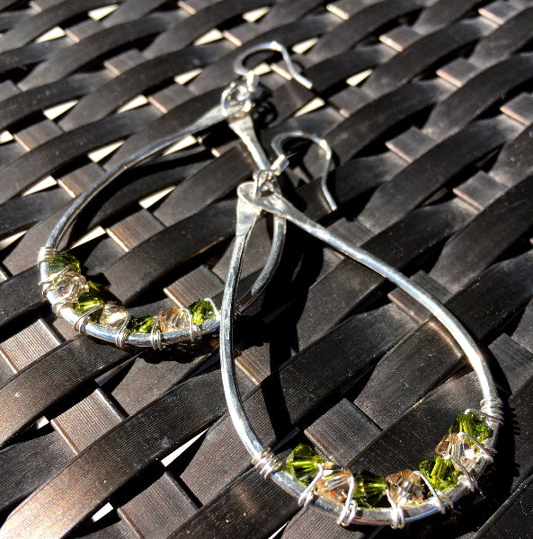 Closeup image of a pair of hammered sterling silver teardrop hoop earrings with silver wire wrap and swarovski crystals on top of a black wicker chair