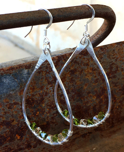 Closeup image of a pair of hammered sterling silver teardrop hoop earrings with silver wire wrap and swarovski crystals hanging from a rustic metal handle