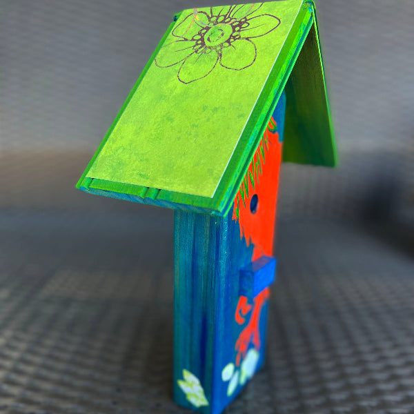 Closeup image of a blue hand painted wooden decorative birdhouse with a green wood roof and decoupaged floral pattern on a wicker chair 