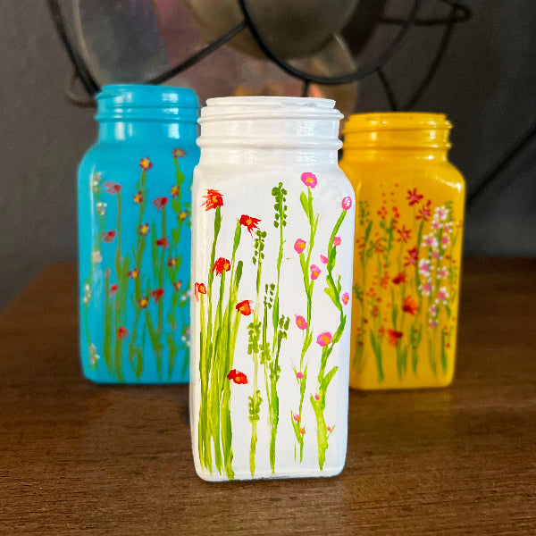 Closeup front view image of three small hand painted bud vases with floral designs painted on front, standing on a wooden table