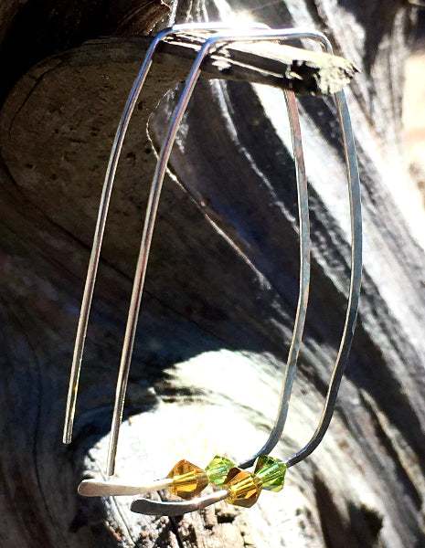 Closeup image of a pair of hammered sterling silver rectangle hoop earrings with swarovski crystals hanging from a piece of wood on the sun