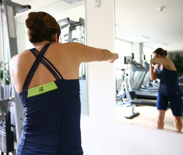 Back view image of a martial arts instructor training in front of a mirror in a fitness center, wearing a navy blue strappy tank top and navy blue long shorts