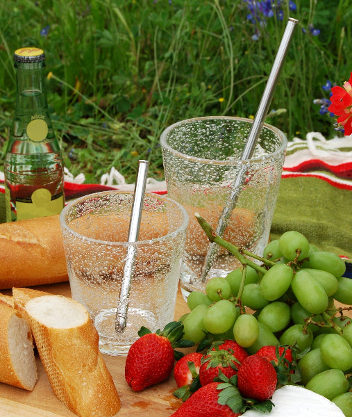 Image of a picnic blanket on the ground in a park with bread, strawberries, green grapes, bottled water and two clear glasses with stainless-steel extendable adjustable metal straws