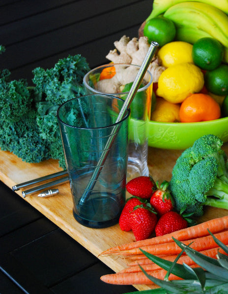 Image of a wooden cutting board with kale, carrots, strawberries, broccoli, and a fruit bowl filled with citrus fruit, bananas and ginger and tall glasses with a stainless-steel extendable adjustable travel straw