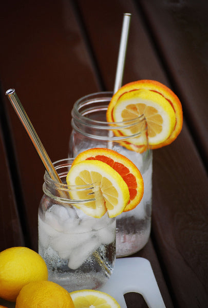 Image of two clear glass mason jars with ice water and sliced citrus fruit and stainless-steel metal extendable sustainable straws on a dark brown wood deck and white cutting board with lemons