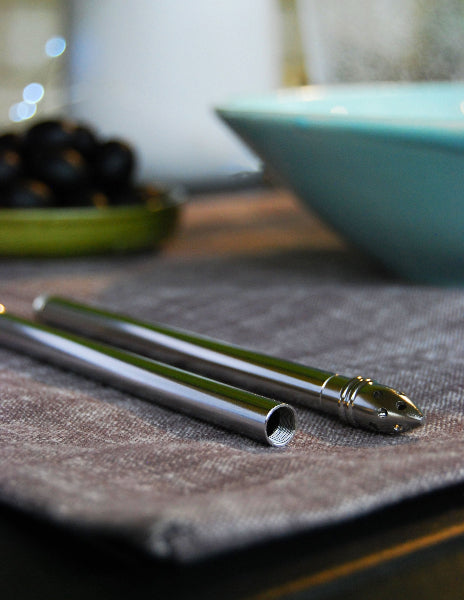 Image of a table setting and a grey woven placemat with a blue bowl on it, a green dish with black olives and a stainless-steel metal extendable travel straw with seed strainer attachment