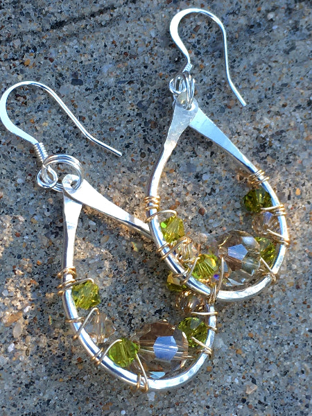 Closeup image of a pair of hammered sterling silver teardrop earrings with gold wire wrap and swarovski crystals on top of a concrete slab