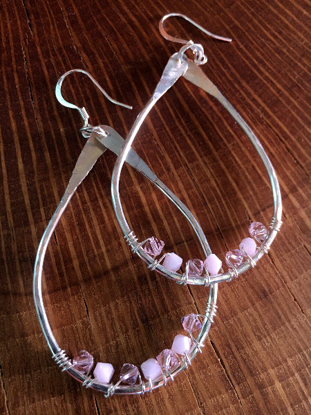 Closeup image of a pair of hammered sterling silver teardrop hoop earrings with silver wire wrap and swarovski crystals laying on a wood stump