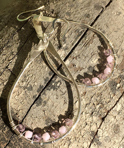 Closeup image of a pair of hammered sterling silver teardrop hoop earrings with silver wire wrap and swarovski crystals displayed ona piece of rustic wood