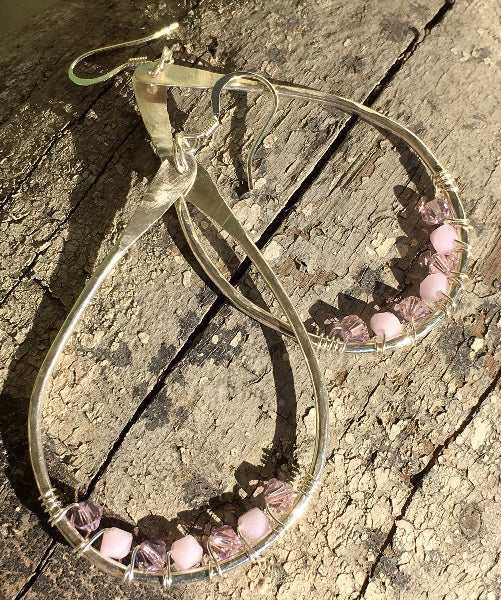 Closeup image of a pair of hammered sterling silver teardrop hoop earrings with silver wire wrap and swarovski crystals displayed ona piece of rustic wood