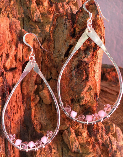 Closeup image of a pair of hammered sterling silver teardrop hoop earrings with silver wire wrap and swarovski crystals hanging from a red stone