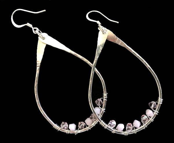 Closeup image of a pair of hammered sterling silver teardrop hoop earrings with silver wire wrap and swarovski crystals against a black background