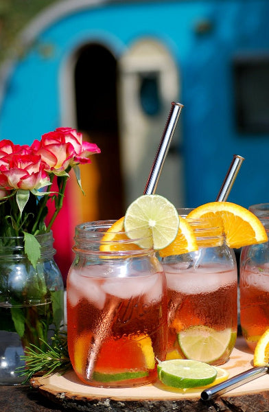 Image of three clear mason jars with sweet tea and citrus slices on the rims and stainless-steel extendable metal straws next to a mason jar with roses and a TaDa turquoise color camper trailer in the background