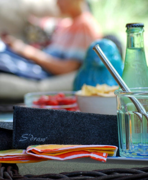 Closeup product image of a black wicker table with snack bowls, a bottle of water, colorful folded napkins, a charcoal color wool travel case with the word S'draw printed on it and a clear glass with a stainless-steel straw